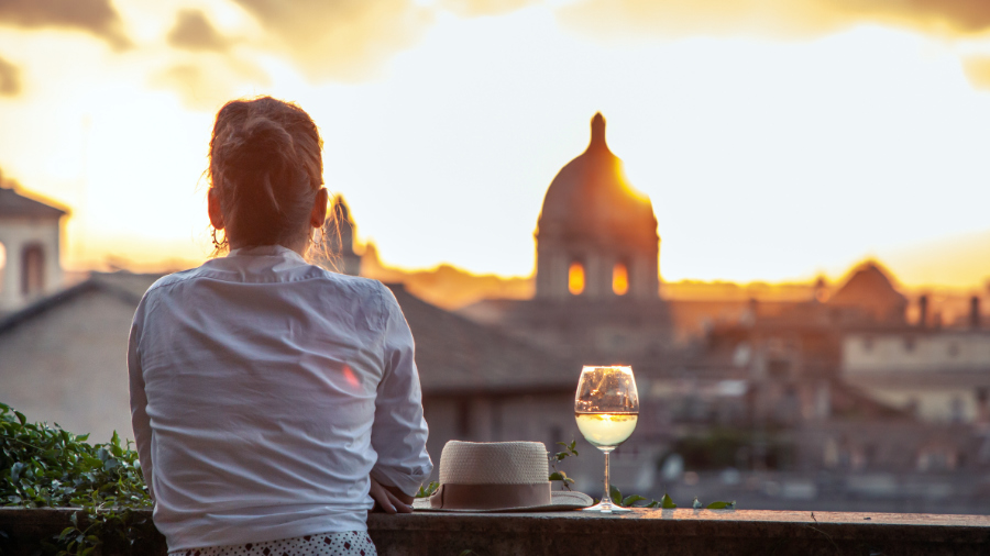 Young woman tourist fashion white dress with glass of white wine in front of panoramic view of Rome cityscape from campidoglio terrace at sunset. Landmarks and domes.